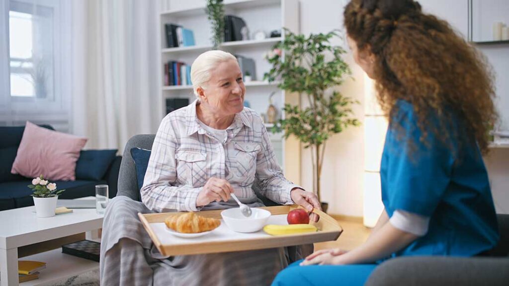 Older couple sits together in an accessible apartment while a caregiver reviews a daily care plan.