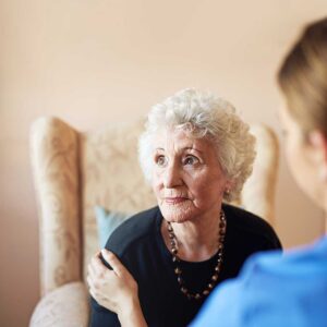 A middle-aged woman sits beside an elderly man in a shared living room, listening attentively and smiling gently.